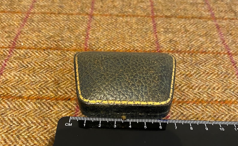 May include: A small, black, leather-bound box with a gold trim. The box is about 6 cm long.