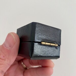 May include: A small, black, square box with a textured surface. The box is held in a hand and has a gold-colored hinge. The box is closed.