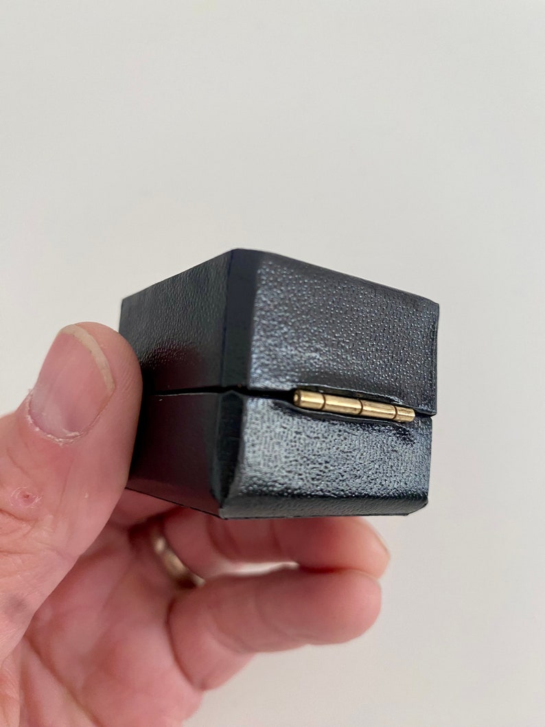 May include: A small, black, square box with a textured surface. The box is held in a hand and has a gold-colored hinge. The box is closed.