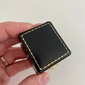 May include: A small, black square box with a gold dotted border and a gold clasp. The box is held in a hand against a white background. The box appears to be made of leather or a similar material.
