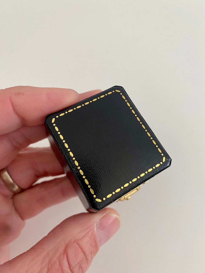 May include: A small, black square box with a gold dotted border and a gold clasp. The box is held in a hand against a white background. The box appears to be made of leather or a similar material.