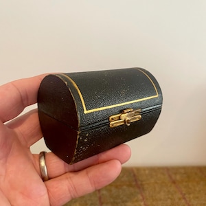 May include: A small, arched, black jewelry box with a textured surface and gold-colored trim. The box features a gold-colored metal latch on the front, which secures the lid. The exterior has visible signs of wear along the edges and corners.