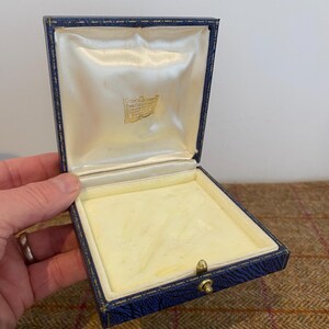 May include: Open blue jewelry box with a cream-colored satin interior and a gold clasp. The box has a square shape and a hinged lid. The interior has a cream-colored velvet lining. The box is empty.