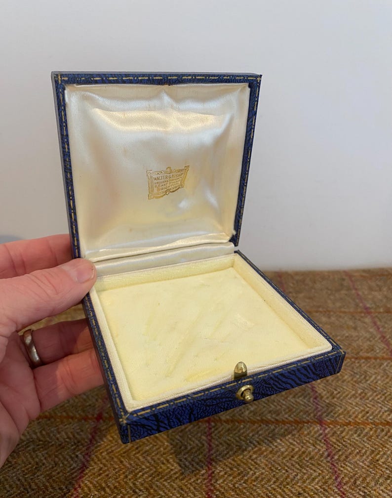 May include: Open blue jewelry box with a cream-colored satin interior and a gold clasp. The box has a square shape and a hinged lid. The interior has a cream-colored velvet lining. The box is empty.