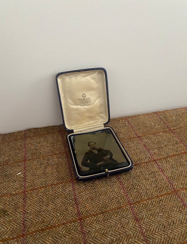 May include: A black and white photograph of a man in a suit and tie, sitting in a chair. The photograph is in a blue velvet case with a white satin lining. The case has a label that reads "W. & D. Downey, Photographers, 107, Regent Street, London, W."