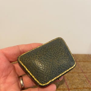May include: A small, dark green leather box with gold trim. The box is closed and has a slightly rounded shape.