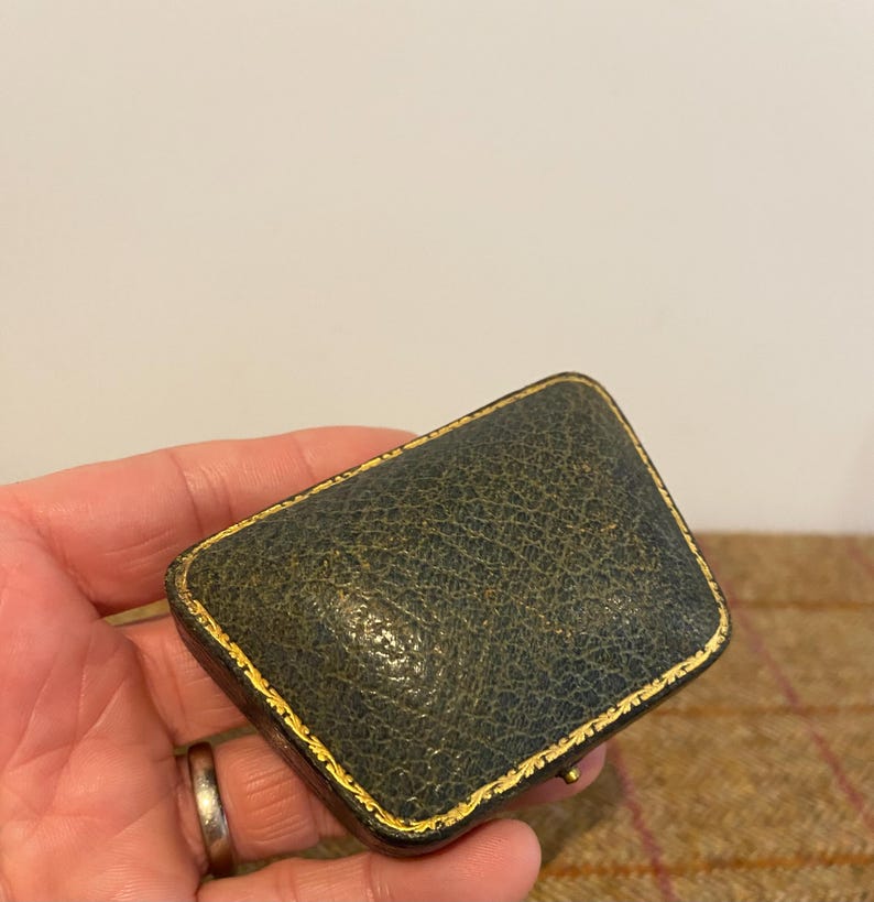 May include: A small, dark green leather box with gold trim. The box is closed and has a slightly rounded shape.