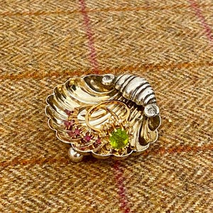 May include: A silver-toned shell-shaped trinket dish featuring a scalloped design and three small ball feet. Inside the dish are two gold rings, one set with a central oval green gemstone surrounded by small clear stones and one with red stones.