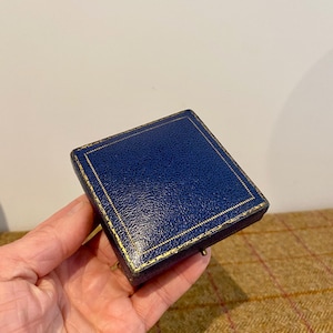 Könnte beinhalten: Eine quadratische, marineblaue Schmuckschatulle mit einer strukturierten, lederartigen Oberfläche. Die Schatulle ist mit goldfarbenen Akzenten versehen und hat einen kleinen Verschluss. Die Schatulle wird in einer Hand gehalten.