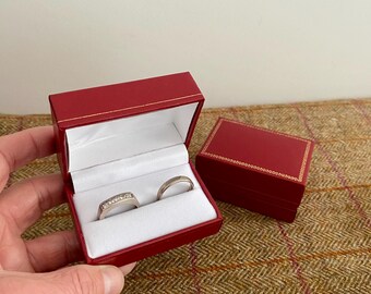 May include: Two wedding bands in a red ring box. The box is open and the rings are visible. The rings are both silver and have small diamonds.
