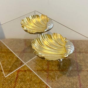 Peut inclure: Deux coupelles métalliques en forme de coquillage avec un intérieur doré poli aux détails nervurés et une charnière argentée contrastante. Chaque coupelle repose sur de petits pieds sphériques, offrant une finition bicolore à bord festonné.