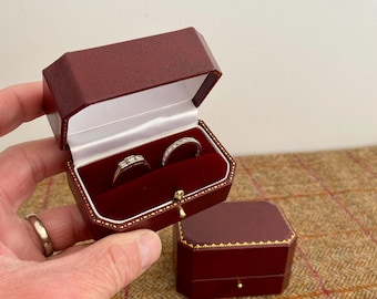 May include: Two rings displayed in a burgundy leather ring box with a gold clasp. The box is open, revealing a white interior and a red velvet lining. A second, closed box is also visible.