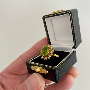 May include: A gold ring with a large oval, light green gemstone, surrounded by smaller clear stones. The ring is displayed in an open black jewelry box with a gold clasp and white interior.