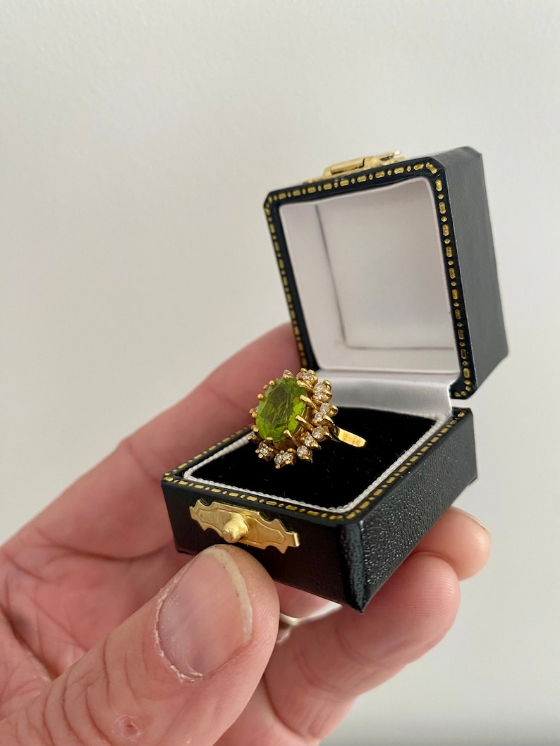 May include: A gold ring with a large oval, light green gemstone, surrounded by smaller clear stones. The ring is displayed in an open black jewelry box with a gold clasp and white interior.