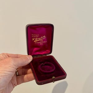 May include: A vintage jewellery box, burgundy in colour, with a bright pink velvet lining. The box is open, revealing a circular recess for a ring or pendant. The interior text reads "Watchmakers & Jewellers H. George & Co. 244 Westminster Bridge S.E."