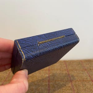 May include: A small, rectangular blue box with a textured pattern and gold trim. The box has a brass hinge and is slightly open, revealing a glimpse of the interior. The box is likely for jewelry or other small items.