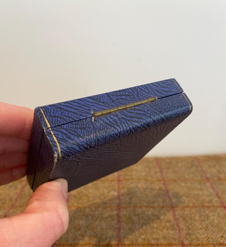 May include: A small, rectangular blue box with a textured pattern and gold trim. The box has a brass hinge and is slightly open, revealing a glimpse of the interior. The box is likely for jewelry or other small items.