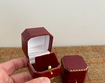 May include: Two red octagonal jewelry boxes. One box is open, revealing a white interior and a red velvet insert. The other box is closed. Both boxes have gold trim and a small gold knob.