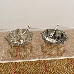 May include: Two silver-toned hexagonal bowls with scalloped edges and intricate floral embossed patterns on the interior. Each bowl includes a matching small silver-toned spoon with a thin handle and a spherical finial resting inside the basin.
