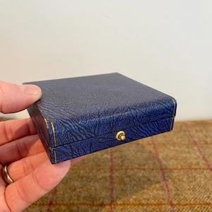 May include: A small, square blue box with a textured pattern and gold trim. The box has a gold-colored clasp. The box is held in a person's hand.