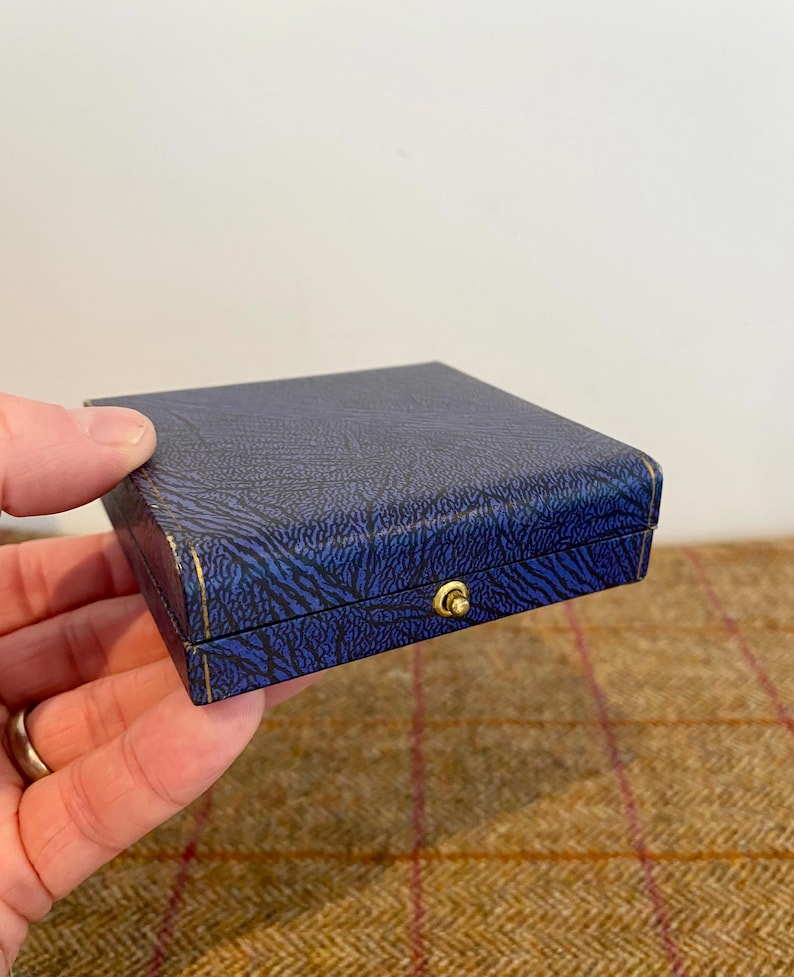 May include: A small, square blue box with a textured pattern and gold trim. The box has a gold-colored clasp. The box is held in a person's hand.