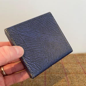 May include: A small, square, navy blue box with a textured surface. The box has gold-colored trim and appears to be closed. The box is held in a hand.