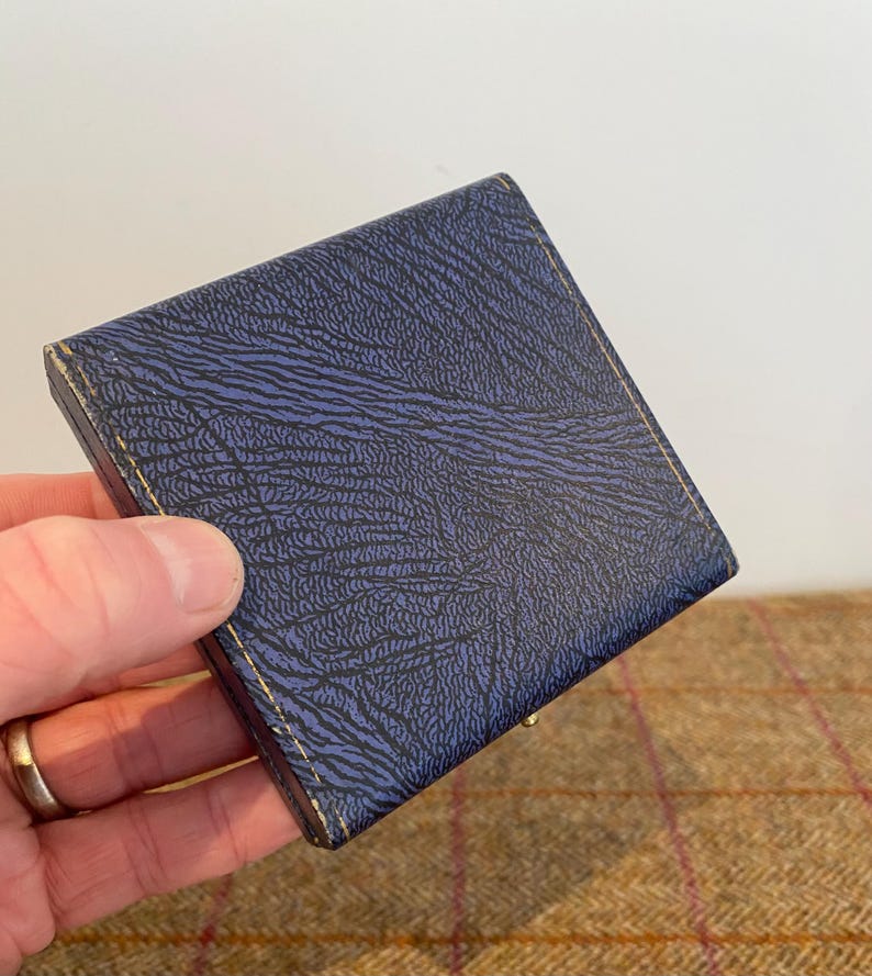 May include: A small, square, navy blue box with a textured surface. The box has gold-colored trim and appears to be closed. The box is held in a hand.