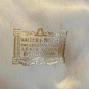 May include: Close-up of a cream-colored satin fabric with a gold-colored rectangular label. The label reads "WALTER G. BISHOP, JEWELLER & SILVERSMITH, 11, EAST STREET, BRIGHTON."