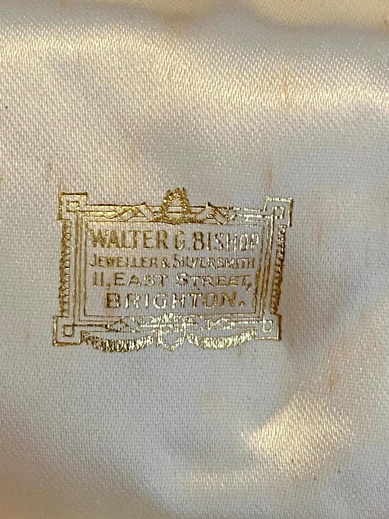 May include: Close-up of a cream-colored satin fabric with a gold-colored rectangular label. The label reads "WALTER G. BISHOP, JEWELLER & SILVERSMITH, 11, EAST STREET, BRIGHTON."
