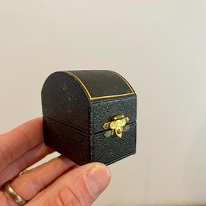 May include: A small, black jewelry box with a curved top and gold trim. The box has a textured surface and a gold-colored clasp. The box is designed to hold small items like rings or earrings.