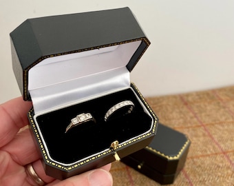 May include: Two rings displayed in a black octagonal jewelry box with a gold trim. One ring features three square stones, and the other is a band with multiple small stones. The box is open, revealing a black velvet interior.