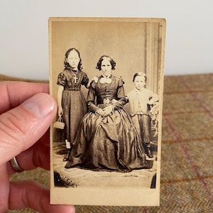 May include: A vintage sepia-toned photograph of a family. A woman in a dark dress sits with two children standing beside her. The girl wears a dress and cross necklace, while the boy is in a suit. The image is held by a hand.