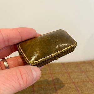 May include: A small, rectangular antique jewellery box. The box is olive green leather with gold trim around the edges. The box is closed with a small clasp. The box is held in a person's hand.