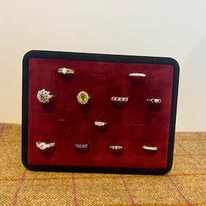 May include: A rectangular black display stand with a deep red velvet surface holding twelve assorted rings. The collection includes rings with various gemstones such as a central oval green stone, red stones, blue stones, and clear white diamonds.
