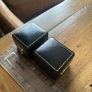 May include: Two black jewelry boxes with gold trim and accents sit on a wooden surface. The boxes are rectangular with hinged lids. A clear ruler with the text "WHSmith" is in the foreground.