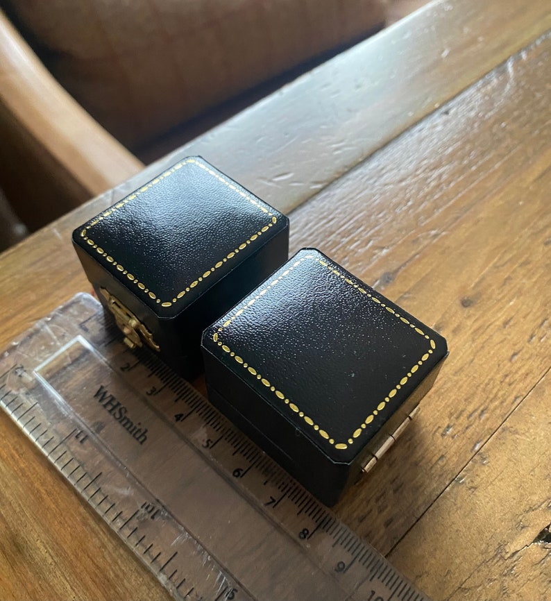 May include: Two black jewelry boxes with gold trim and accents sit on a wooden surface. The boxes are rectangular with hinged lids. A clear ruler with the text "WHSmith" is in the foreground.