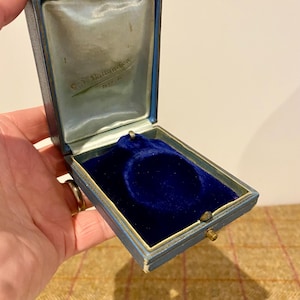 Può includere: Scatola portagioie blu vintage aperta, con interno in velluto. La scatola ha un esterno azzurro con finiture dorate e una chiusura in ottone. L'interno è foderato con tessuto di raso azzurro e una base in velluto blu scuro. Il testo "S.E. Ballantyne, N.Y." è visibile.