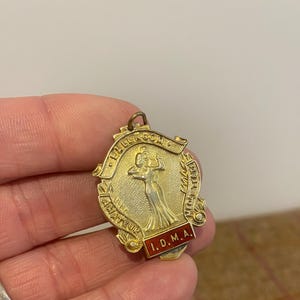 Könnte beinhalten: Goldene Medaille mit einem rot-weißen Band. Die Medaille zeigt eine Frau in einem Kleid, die eine Trophäe hält. Der Text auf der Medaille lautet "Ballroom Amateur First Prize I.D.M.A."