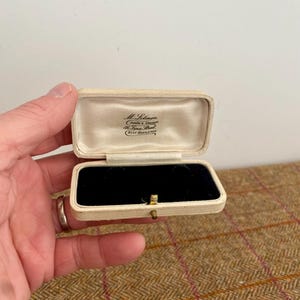 Könnte beinhalten: Eine weiße, rechteckige Schmuckschatulle mit einem Scharnierdeckel. Der Deckel ist geöffnet und zeigt ein Etikett im Inneren mit der Aufschrift "M. Levinger, Juwelier & Silberschmied, 81, Town Street, West Bridgford". Die Schachtel hat ein schwarzes Samt-Innenfutter mit drei Fächern.