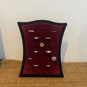May include: A black display stand with a deep red velvet surface holding twelve assorted rings. The rings feature various metal bands, including silver and gold tones, set with clear, green, and dark gemstones in diverse shapes and sizes.