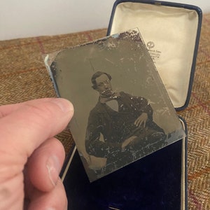 May include: A black and white tintype photograph of a man in a dark suit and a white shirt with a bow tie. The photograph is in a blue velvet case.