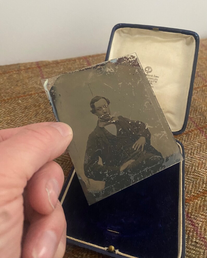 May include: A black and white tintype photograph of a man in a dark suit and a white shirt with a bow tie. The photograph is in a blue velvet case.