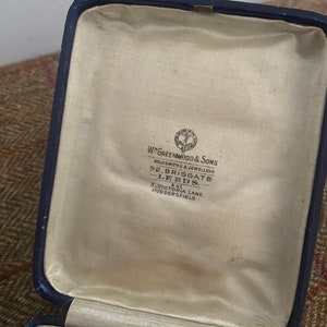 May include: A blue velvet jewelry box with a cream colored lining. The inside of the box has a label that reads "W. Greenwood & Sons Goldsmiths & Jewellers 92, Briggate Leeds. 3, Victoria Lane. Huddersfield."