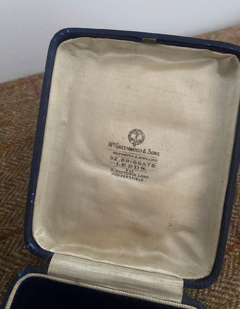 May include: A blue velvet jewelry box with a cream colored lining. The inside of the box has a label that reads "W. Greenwood & Sons Goldsmiths & Jewellers 92, Briggate Leeds. 3, Victoria Lane. Huddersfield."