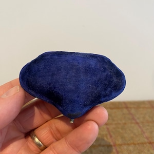 May include: A dark blue velvet jewelry box with an unusual, organic shape. The box is held in a hand, highlighting its rich colour and texture. The box has a small metal post on the bottom.