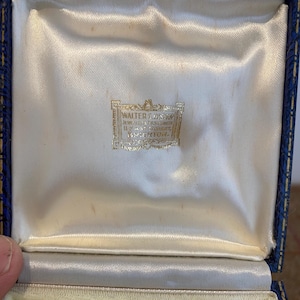 May include: An open, vintage jewelry box with a blue exterior and a cream-colored satin interior. The box features a gold-colored label with the text "WALTER COUSINS, JEWELLER & GOLDSMITH, 11, EAST STREET, BRIGHTON."