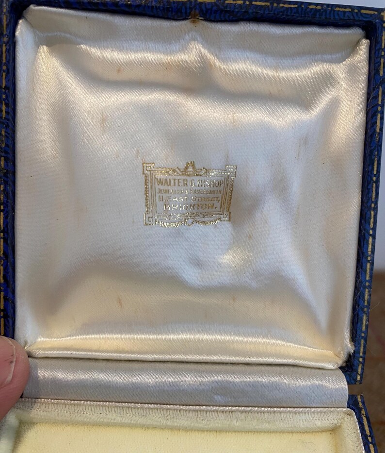 May include: An open, vintage jewelry box with a blue exterior and a cream-colored satin interior. The box features a gold-colored label with the text "WALTER COUSINS, JEWELLER & GOLDSMITH, 11, EAST STREET, BRIGHTON."