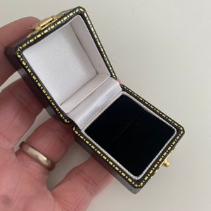 May include: An open, black leatherette jewelry box with a gold clasp and trim. The interior features a white satin lining on the lid and a black velvet base. The box is designed to hold a ring or other small jewelry items.
