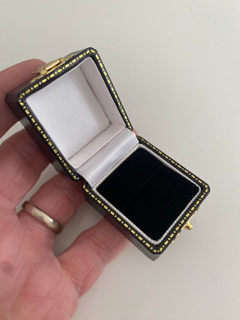May include: An open, black leatherette jewelry box with a gold clasp and trim. The interior features a white satin lining on the lid and a black velvet base. The box is designed to hold a ring or other small jewelry items.