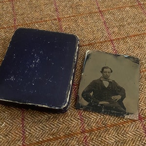 May include: A black and white tintype photograph of a man wearing a suit and tie. The photograph is in a blue velvet case.